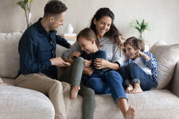 Laughing 30s couple tickling little preschool kids seated on sofa in living room. Hispanic parents enjoy playtime with adorable son and cute daughter. Games of happy family at home, love, fun concept - obrazy, fototapety, plakaty