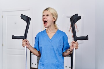 Young physiotherapist woman holding crutches angry and mad screaming frustrated and furious,...