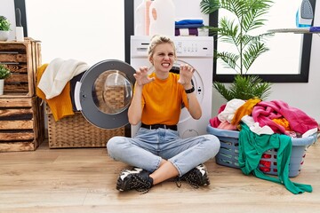 Young blonde woman doing laundry sitting by washing machine smiling funny doing claw gesture as...