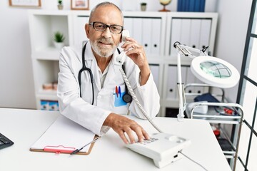 Senior grey-haired man wearing doctor uniform talking on the telephone at clinic
