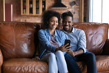 Obraz premium Happy millennial African American family couple using cellphone, posing together for selfie photo or recording video on web camera, holding distant online conversation, modern tech addiction.