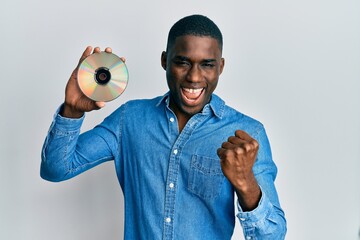 Young african american man holding compact disc screaming proud, celebrating victory and success...