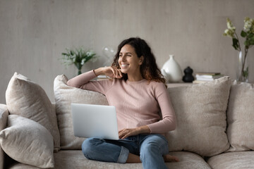 Happy dreamy woman with laptop sitting on cozy couch, smiling excited pensive beautiful young female looking to aside, distracted from online chatting, visualizing, enjoying leisure time with device