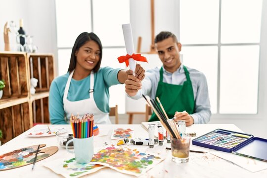 Young latin painter couple smiling happy holding diploma sitting on the table at art studio