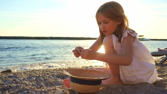 girl in white dress sits on seashore and collects pebbles and twigs in straw hat