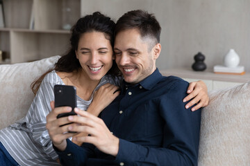 35s Latin couple sit on sofa watch funny videos on cellphone, wife and husband relaxing indoors laughing feels carefree spend weekend with modern wireless tech, online amusing content, fun concept