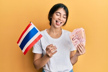 Young caucasian woman holding thailand flag and baht banknotes sticking tongue out happy with funny...
