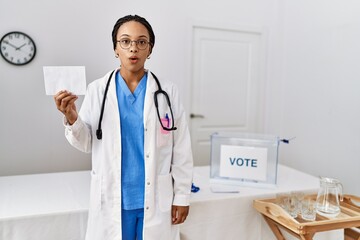 Young african american doctor woman voting holding envelope scared and amazed with open mouth for...