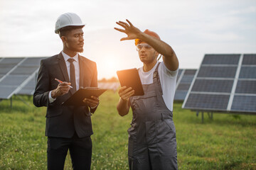 Indian engineer in yellow hardhat and safety glasses holding digital tablet and talking with male owner of solar plant. African american man in suit checking production of alternative energy.