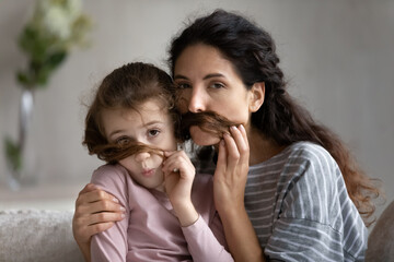 Headshot portrait of mother with cute daughter having fun with hair, making funny faces with moustache together, happy young mom and adorable little kid looking at camera, posing for family photo