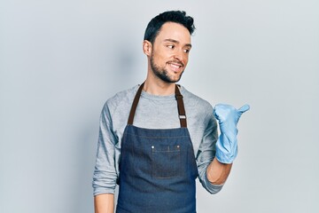 Young hispanic man wearing apron and gloves smiling with happy face looking and pointing to the...