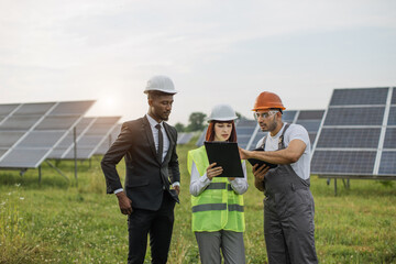 Male and female inspectors in white helmets talking with technician among solar panels outdoors. Multiracial people examining and maintaining production of alternative energy.