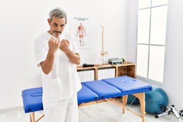 Middle age hispanic therapist man working at pain recovery clinic ready to fight with fist defense gesture, angry and upset face, afraid of problem