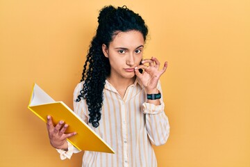 Young hispanic woman with curly hair holding book mouth and lips shut as zip with fingers. secret...