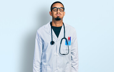Fototapeta premium Young african american man wearing doctor uniform and stethoscope puffing cheeks with funny face. mouth inflated with air, crazy expression.