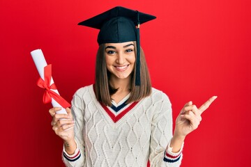 Young brunette girl holding graduate degree diploma smiling happy pointing with hand and finger to the side - obrazy, fototapety, plakaty