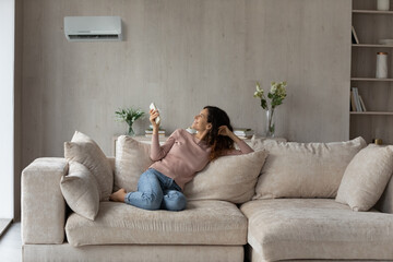 Smiling woman using air conditioner remote controller, relaxing on comfortable couch in living room...
