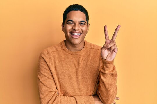 Young handsome hispanic man wearing casual winter sweater smiling with happy face winking at the camera doing victory sign. number two.