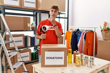 Young caucasian volunteer man packing donations box at charity center serious face thinking about...