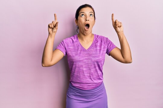 Young latin woman wearing sportswear amazed and surprised looking up and pointing with fingers and raised arms.