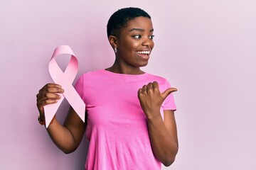 Young african american woman holding pink cancer ribbon pointing thumb up to the side smiling happy with open mouth