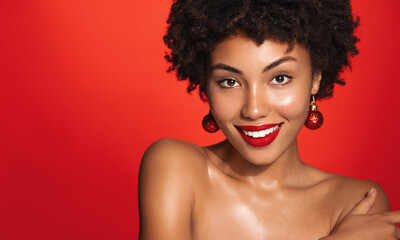 Portrait of beautiful Black woman on red background, red lipstick and festive makeup, Christmas tree earrings, glowing clean body and skin, Winter holidays concept