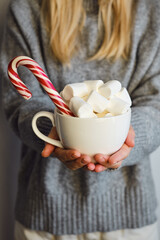 The girl holds in her hands a cup of cocoa with marshmallows and caramel canes. Hot chocolate drink and sweets. New Year and Christmas festive mood, festive home cozy decor
