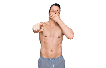 Handsome young man wearing swimwear shirtless laughing at you, pointing finger to the camera with hand over mouth, shame expression