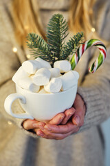 The girl holds in her hands a cup of cocoa with marshmallows and caramel canes. Hot chocolate drink and sweets. New Year and Christmas festive mood, festive home cozy decor
