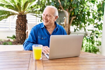 Senior caucasian man using laptop and drinking orange juice sitting on the table at terrace.