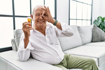 Senior man holding pills doing ok gesture with hand smiling, eye looking through fingers with happy face.