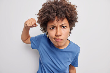 Photo of outraged angry woman clenches fist and looks irritated frowns face doesnt like somethning wears casual blue t shirt isolated over white background. Irritated female model feels rage