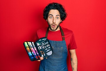 Handsome hispanic man wearing apron holding sew kit scared and amazed with open mouth for surprise,...