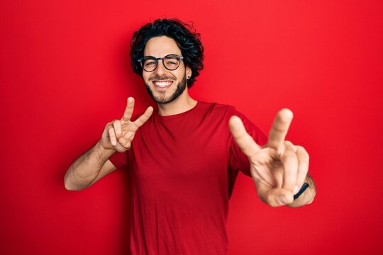 Handsome hispanic man wearing casual t shirt and glasses smiling looking to the camera showing fingers doing victory sign. number two.