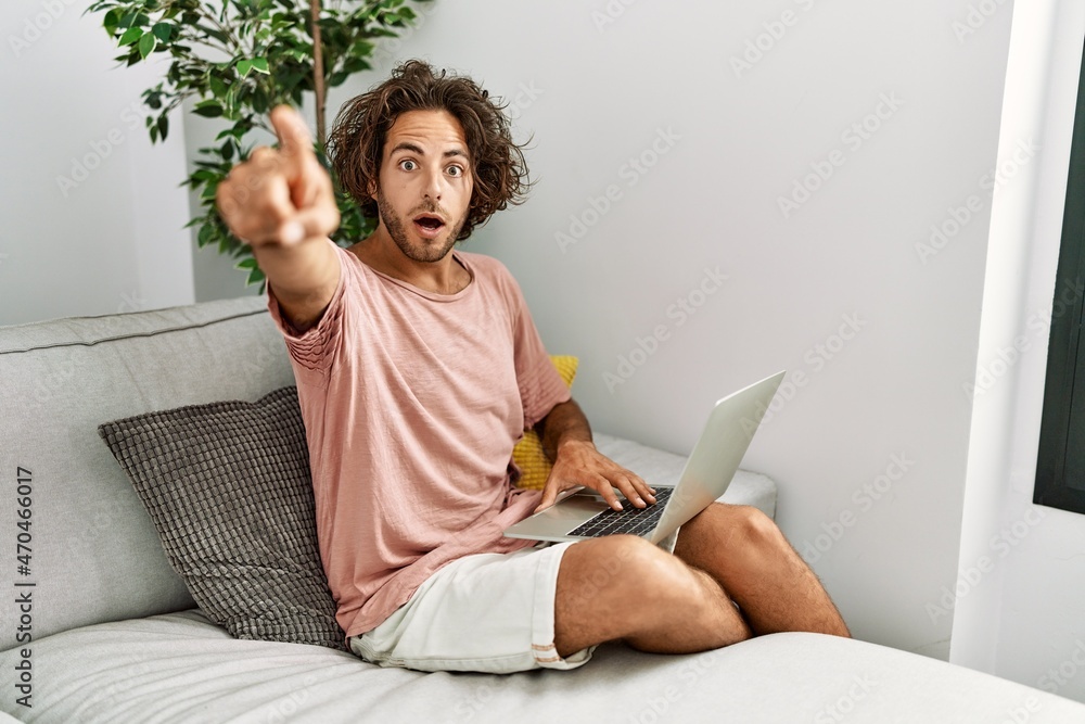 Sticker young hispanic man sitting on the sofa at home using laptop pointing with finger surprised ahead, op