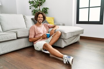 Young hispanic man drinking coffee sitting on the floor at home.