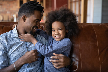 Obraz premium Affectionate happy loving young African American father tickling cute little multiracial kid daughter, playing on weekend, having fun together on comfortable sofa, good family relations concept.