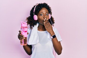 Young african american woman wearing sportswear holding water bottle using headphones covering...