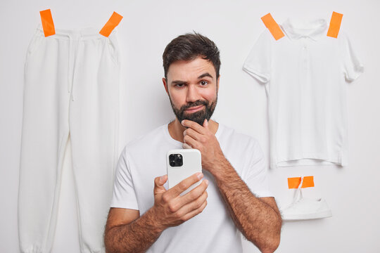 Serious Confident Man Raises Eyebrows Holds Chin And Looks Directly At Camera Holds Mobile Phone Poses Against White Background With Trousers T Shirt And Sneaker Thinks How To Sell Clothes Online.