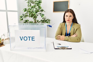 Young brunette woman sitting at election table with voting ballot skeptic and nervous, frowning...