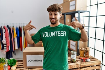 Young arab man wearing volunteer t shirt at donations stand looking confident with smile on face, pointing oneself with fingers proud and happy.