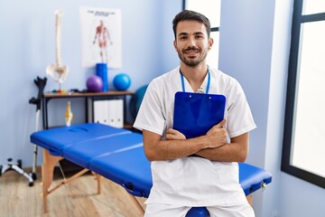 Young hispanic man wearing physiotherapist uniform holding clipboard at rehab clinic