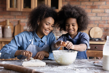 Smiling young African ethnicity mother nanny watching little funny multiracial child girl adding eggs to flour, involved in preparing homemade pastry together on weekend in kitchen, hobby concept. - obrazy, fototapety, plakaty