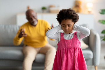 Sad black little girl closes her ears and does not want to listen to swear old man in living room