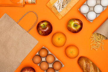 Paper eco bag, bottle of olive oil, eggs, bread, beans, red apples, fruits, spiral paste on orange background