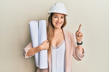 Young blonde woman wearing safety helmet holding blueprints smiling happy pointing with hand and...
