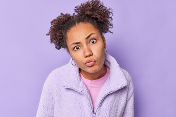 Curly haired beautiful young woman looks with attentive gaze at camera pouts lips focused at something dressed in warm fur jacket listens information attentively isolated over purple background.