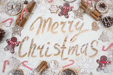 The inscription on the flour Merry Christmas. A frame of gingerbread men, cinnamon, cookies, lollipops and Christmas decorations on a wooden background. The concept of cafes and restaurants. 