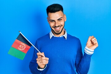 Young hispanic man with beard holding afghanistan flag screaming proud, celebrating victory and...