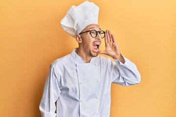 Bald man with beard wearing professional cook uniform shouting and screaming loud to side with hand...